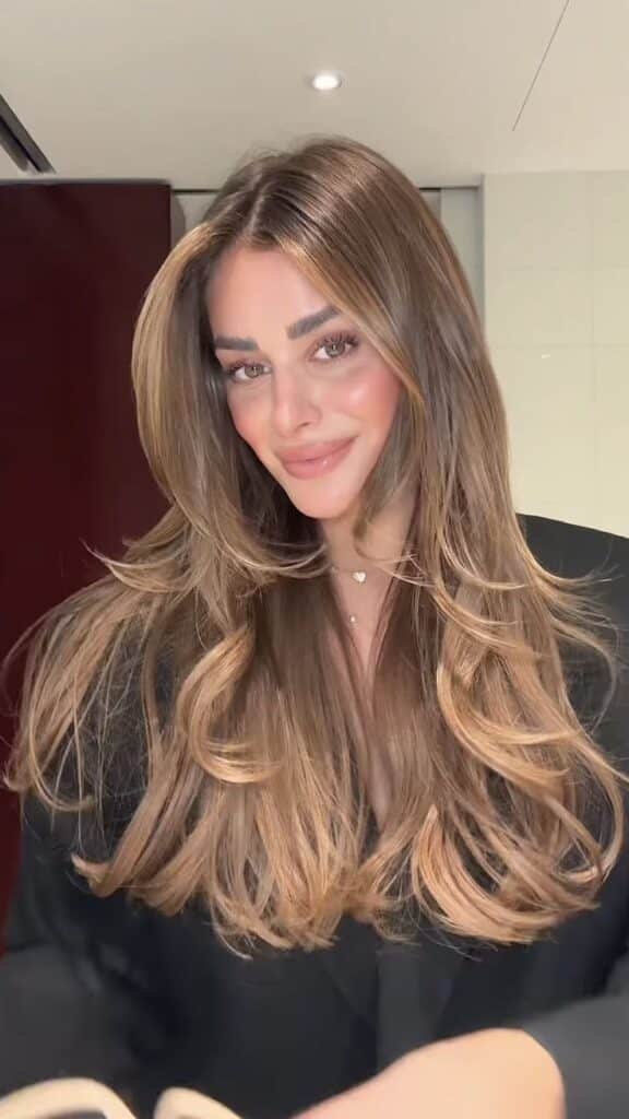 Smiling woman with long, wavy brown hair wearing a black outfit in a brightly lit room.