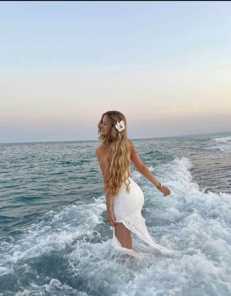 Woman in white dress with flower in hair standing in ocean waves at sunset.