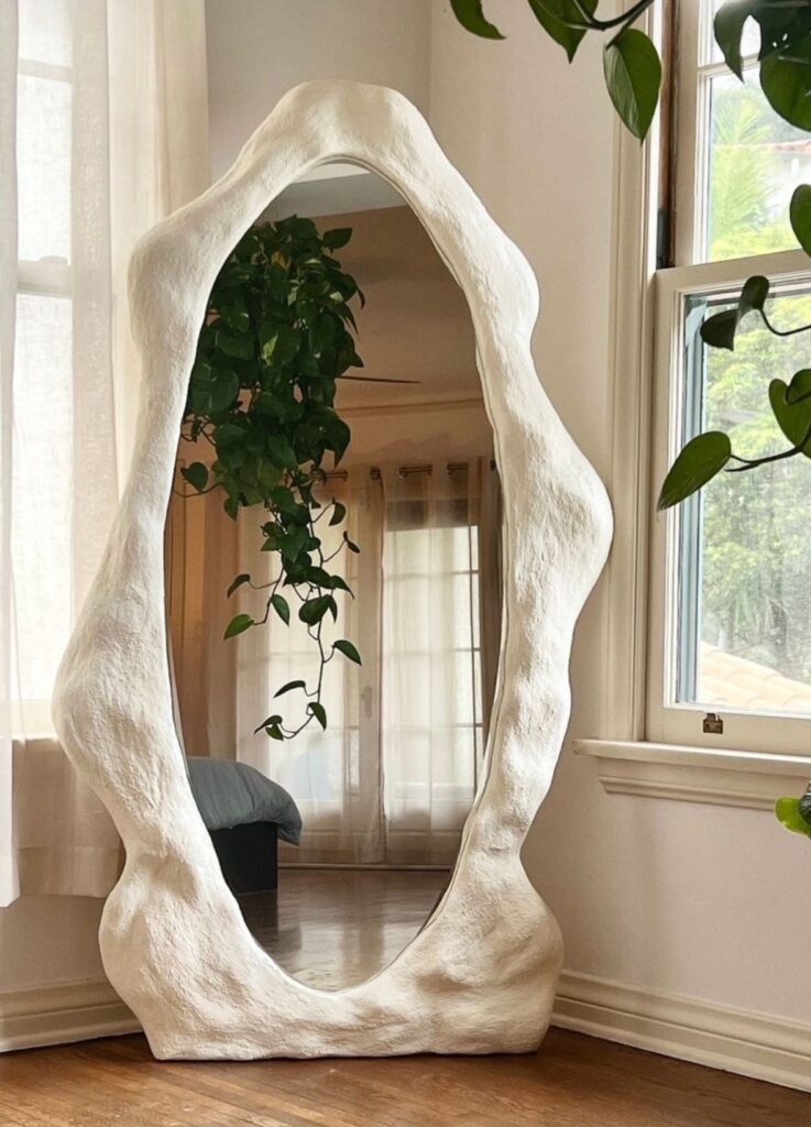 Unique sculptural mirror with abstract frame reflecting greenery in a cozy, well-lit room.