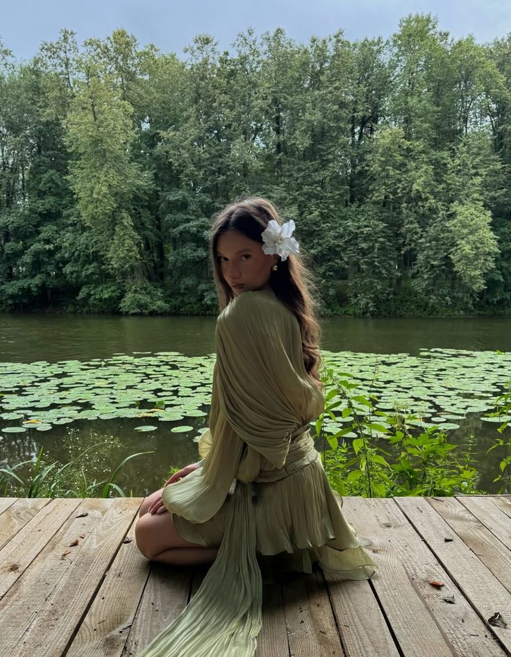 Woman in green dress with flower in hair sits by a serene pond with lily pads and lush forest backdrop.