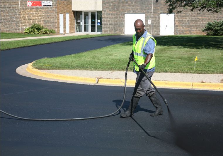 Man in high-visibility vest sealcoating a parking lot on a sunny day.