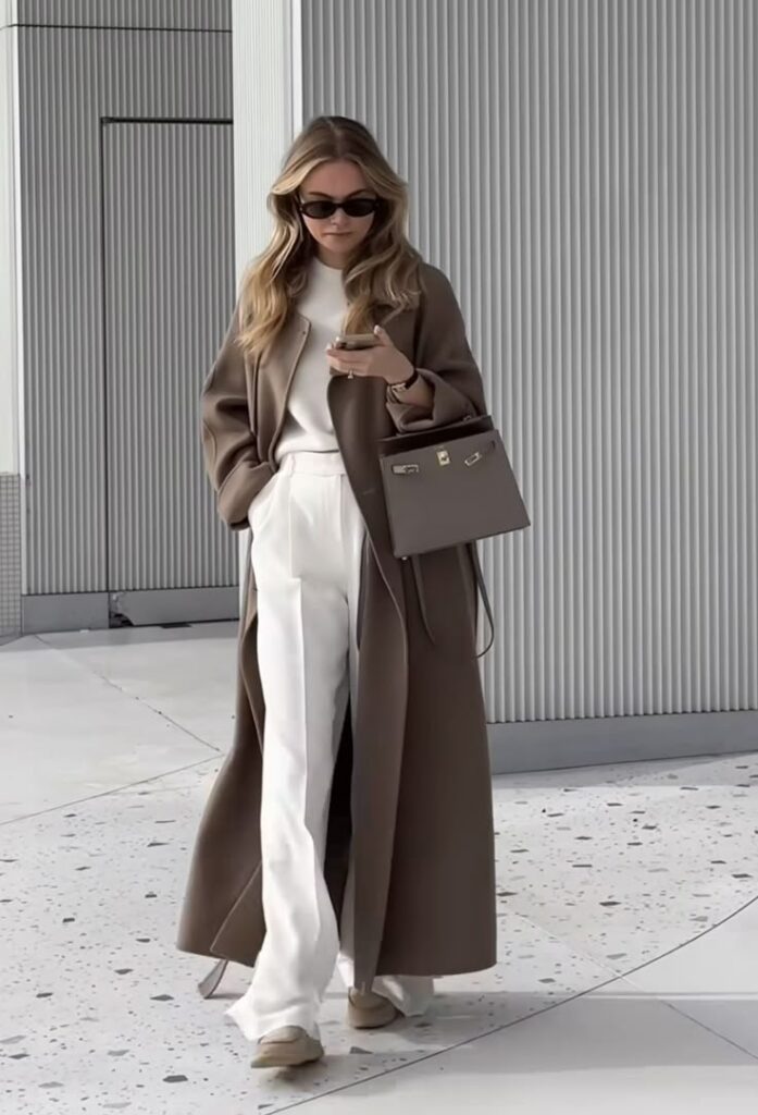 Stylish woman in brown coat and white pants, carrying handbag, using phone, walking urban street. Fashionable streetwear.