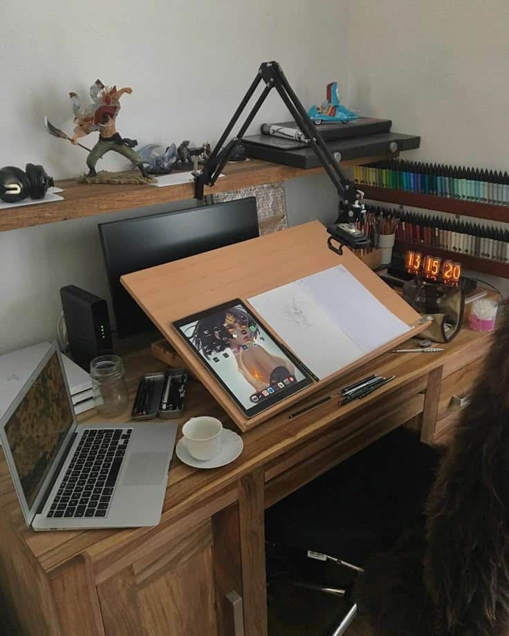 Artist's workspace with drawing setup, laptop, tablet, colorful pens, and coffee cup on a wooden desk.