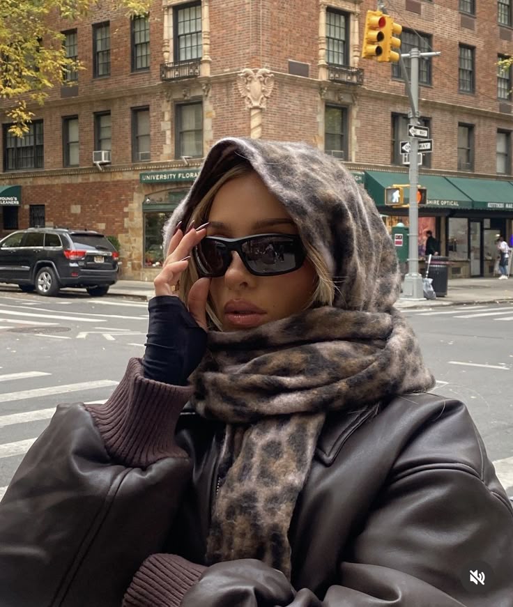 Woman in stylish winter outfit with sunglasses and scarf, standing at a city intersection with brick buildings in the background.