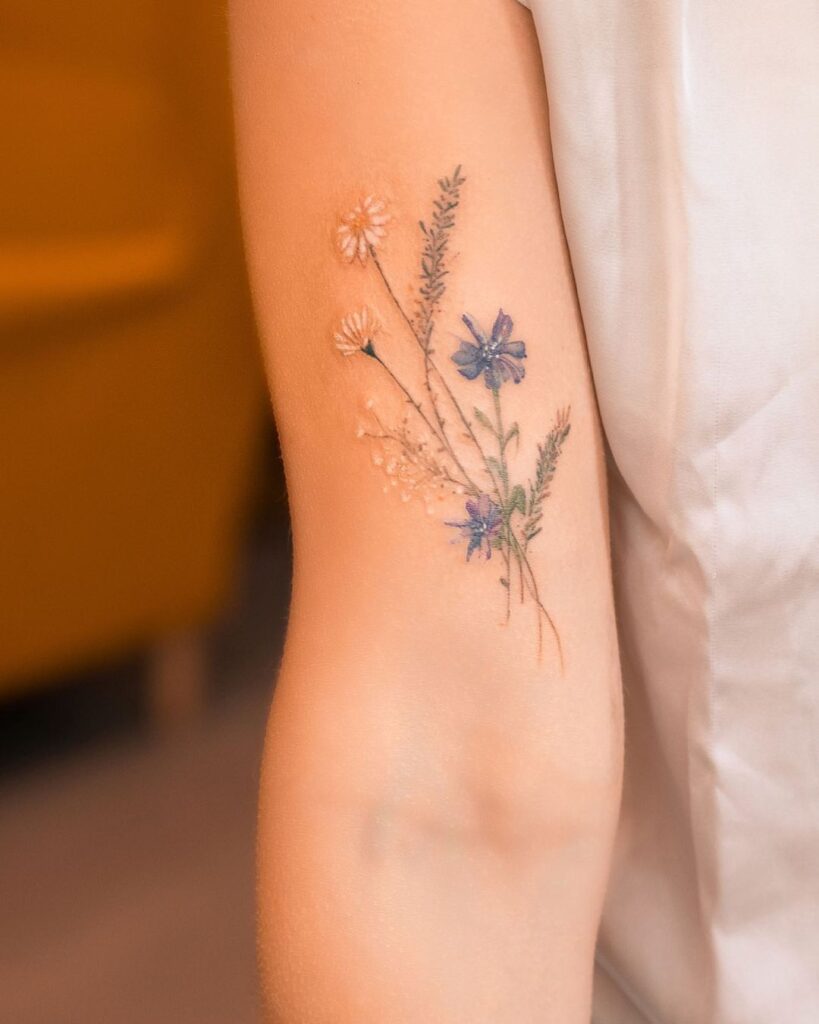 Delicate floral tattoo on a person's inner arm, featuring purple and white flowers with green foliage.