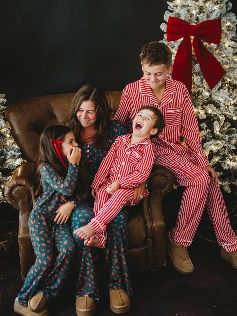 Family in matching holiday pajamas, sitting on a couch by a Christmas tree with lights and festive decor.