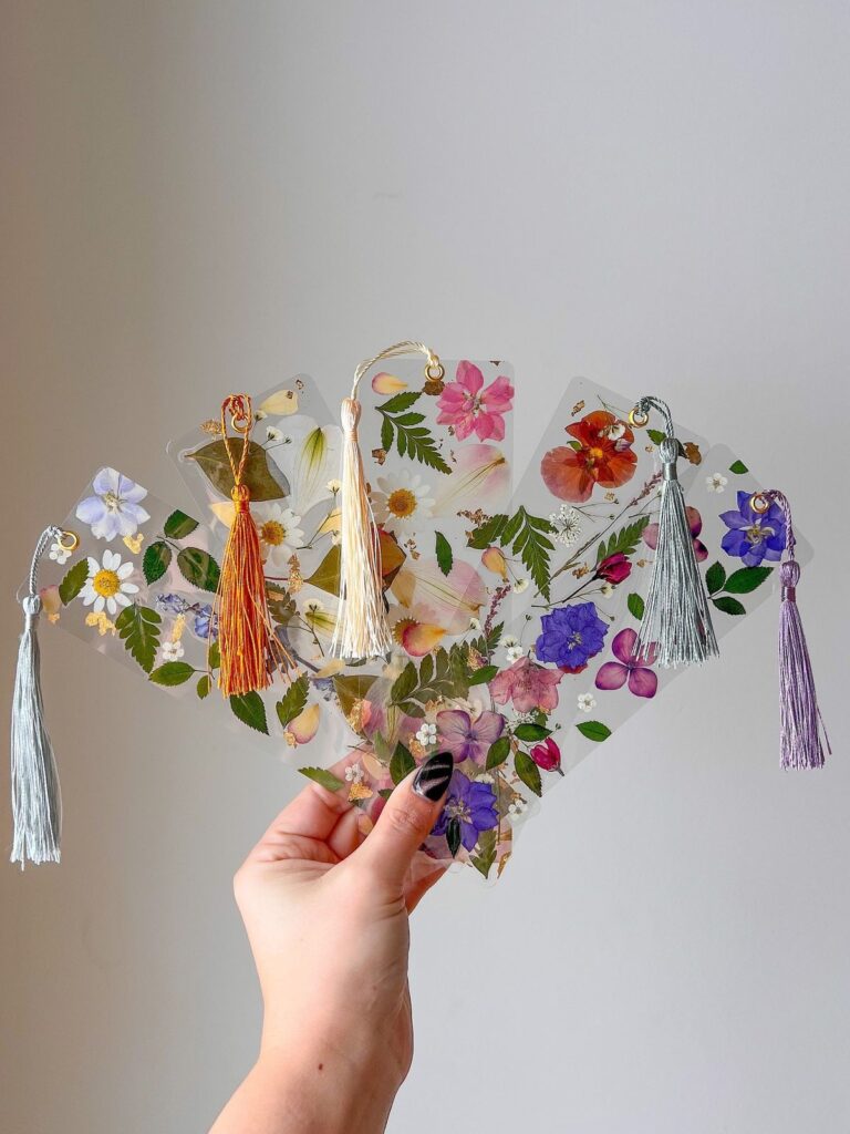 Hand holding floral bookmarks with tassels, featuring colorful pressed flowers for creative and decorative reading accessories.