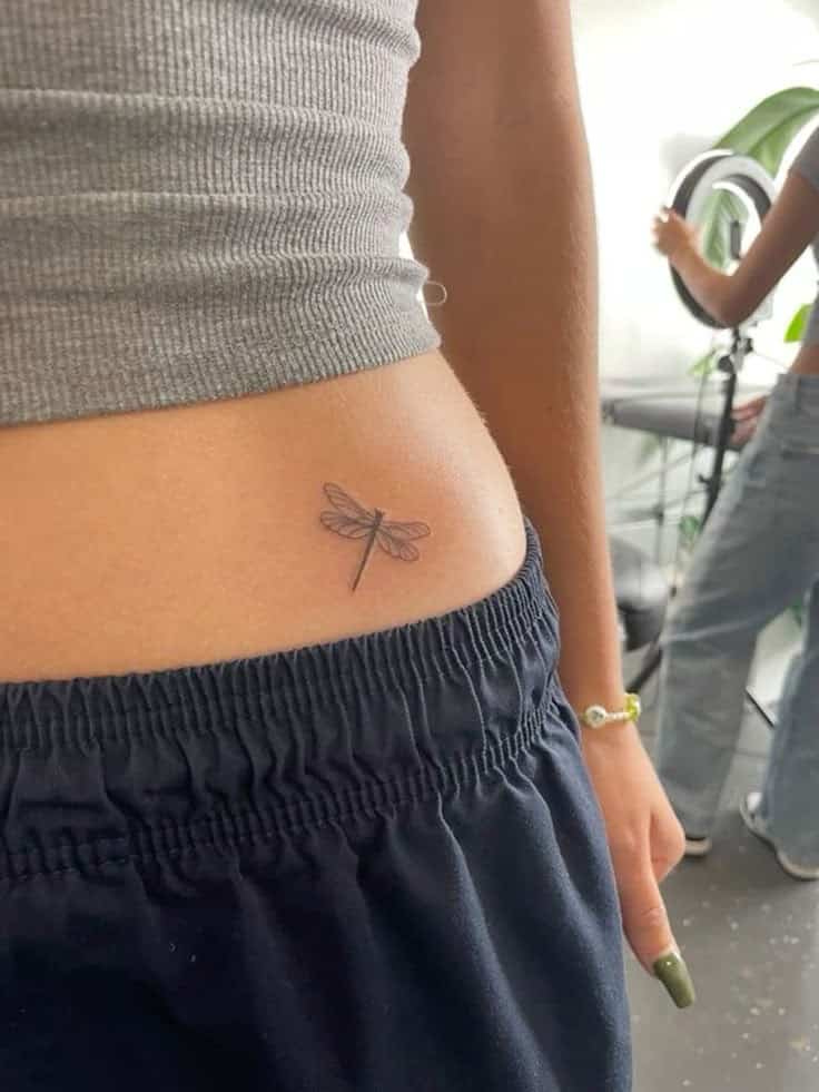 Dragonfly tattoo on a person's waist, seen close up, wearing a gray top and dark pants.