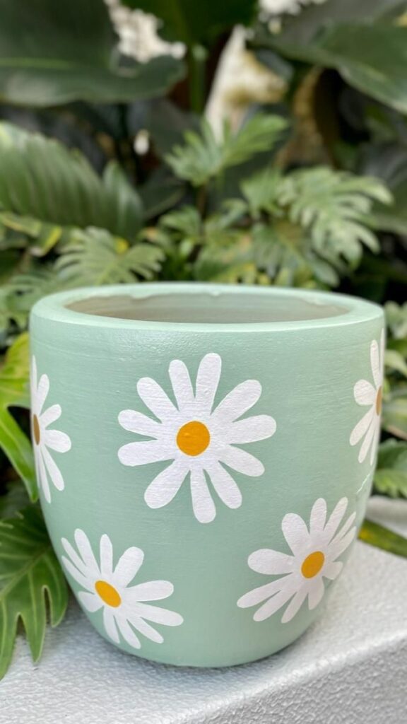 Green flower pot with white daisy design, set against lush greenery. Perfect for garden decor or plant display.
