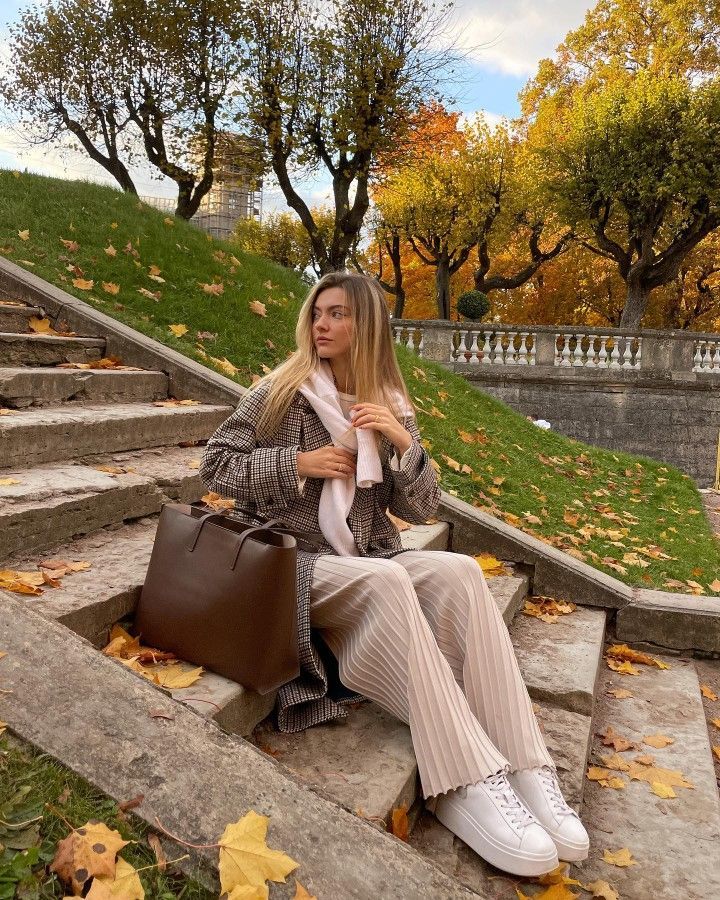 Woman sitting on autumn steps, wearing a coat and scarf, with a brown bag, surrounded by colorful leaves and trees.