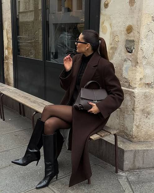 Stylish woman in long coat and boots sitting on bench, holding handbag and wearing sunglasses.