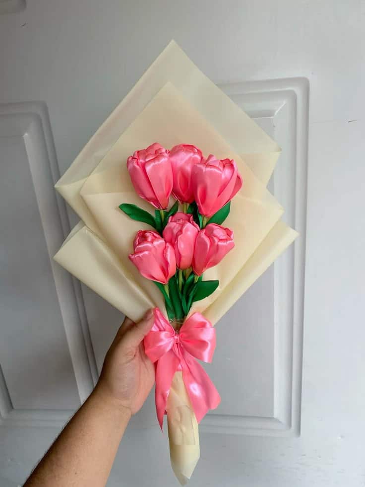 Hand holding a bouquet of pink tulips with green leaves, wrapped in cream paper and tied with a pink ribbon bow.