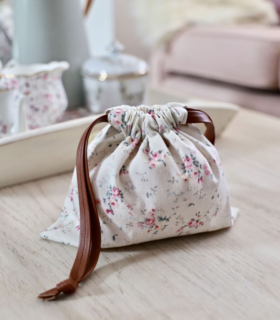 Floral drawstring bag with a leather strap on a wooden table, near a soft-focus vintage tea set.