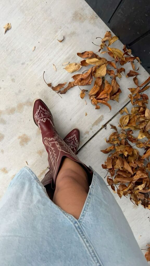 Woman wearing red cowboy boots standing among fallen leaves on a sidewalk.