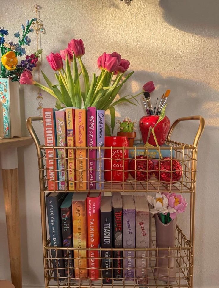 Bookshelf with colorful books, pink tulips, and art supplies in a golden cart, creating a cozy and creative atmosphere.