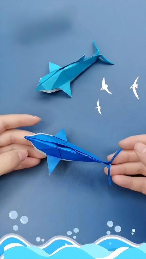 d5b494e8e28a1a4c9344063795b4e474 Hands holding blue origami dolphin and shark on ocean-themed background. | Sky Rye Design Hands holding blue origami dolphin and shark on ocean-themed background.