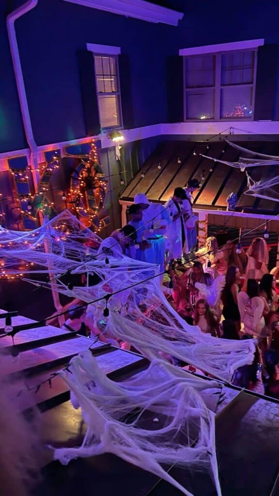 Halloween party with decorations, colorful lights, cobwebs, and people in costumes in a festive atmosphere.