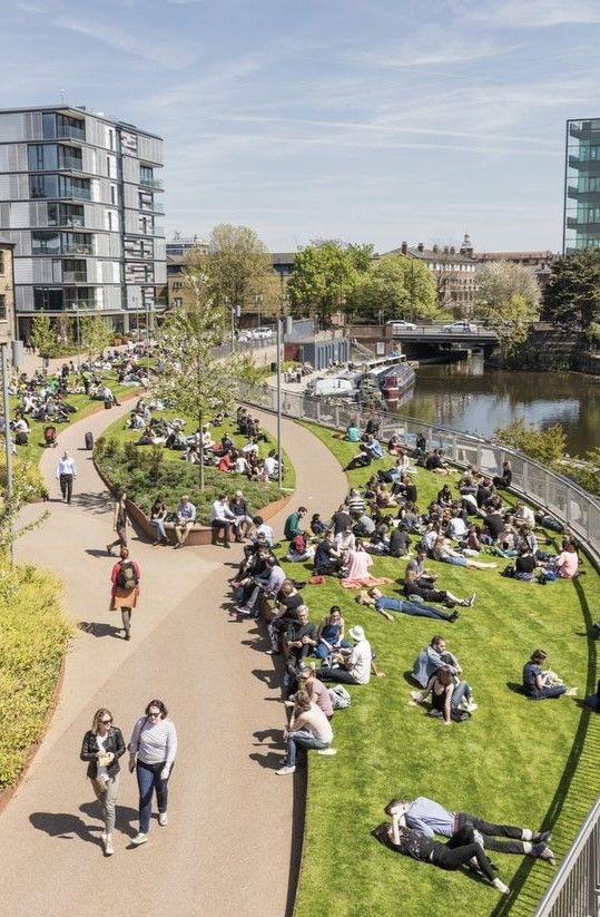 People relaxing in a riverside park on a sunny day, surrounded by modern buildings and greenery.