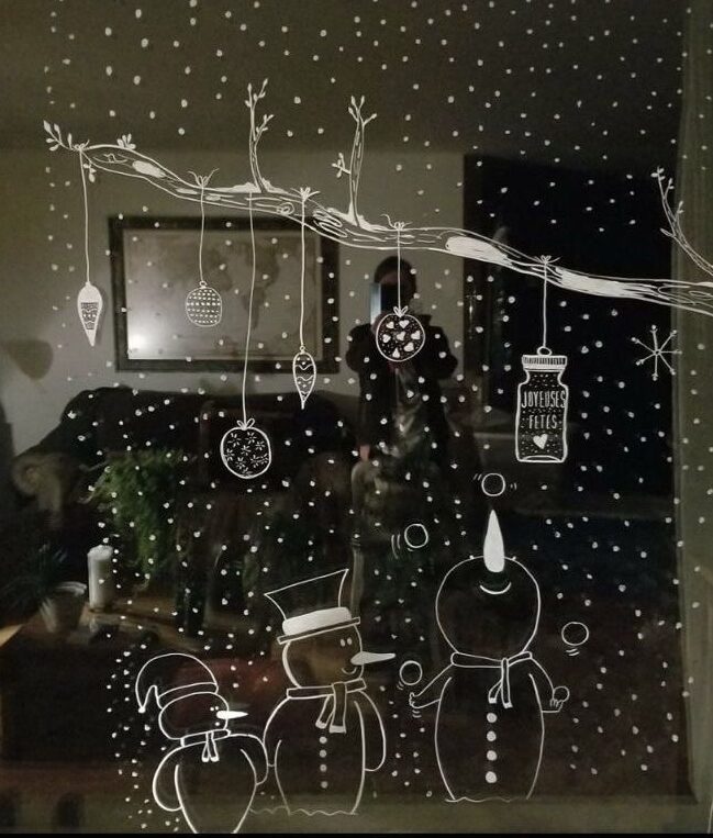 Festive window art with snowflakes, ornaments, and snowmen drawing, reflecting cozy living room setting.