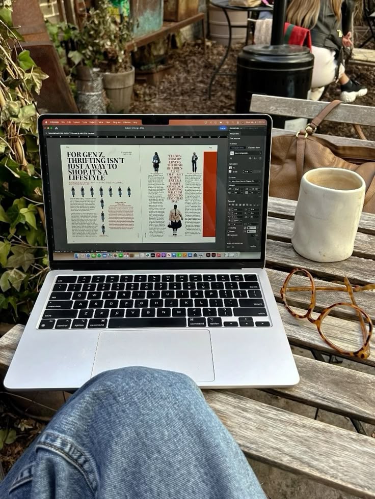 Laptop showing design work on a wooden table with coffee and glasses, outdoor workspace environment.