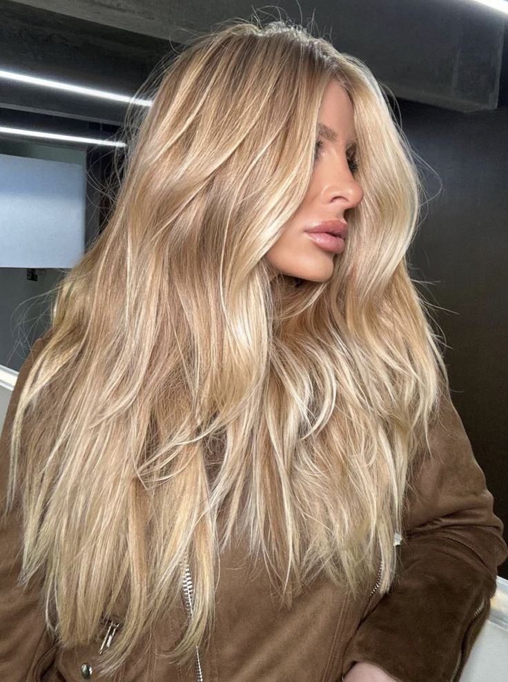 Blonde woman with long, wavy hair wearing a brown jacket in a modern, indoor setting.