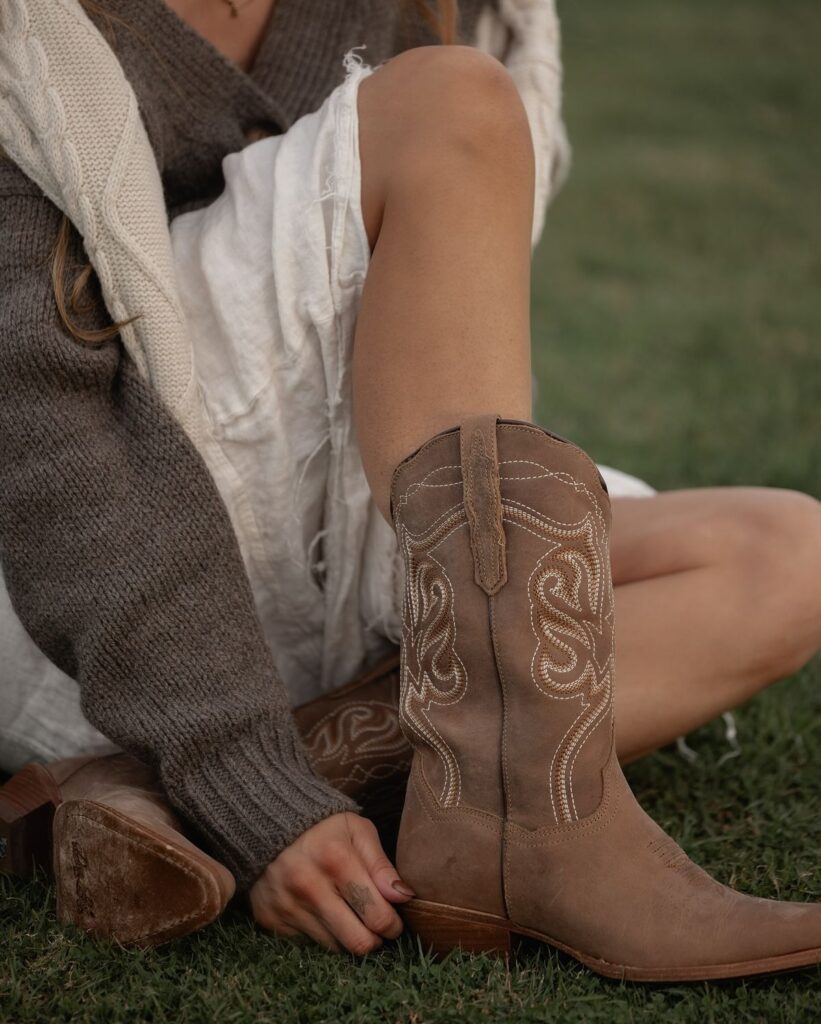 Close-up of a person wearing brown cowboy boots and a white skirt, sitting on grass with a brown sweater.