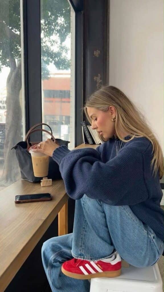 Woman in cozy sweater sitting by window, holding iced coffee, sneakers visible. Casual cafe vibe.