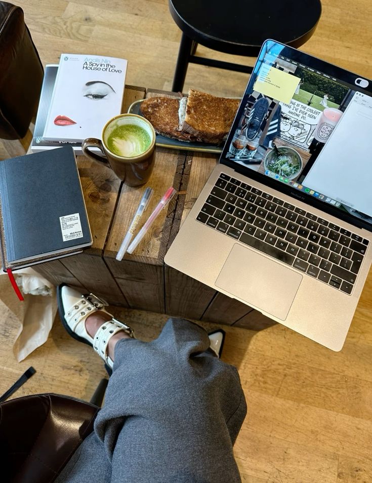 Cozy workspace with laptop, books, matcha latte, and toast on a wooden table in a cafe setting.