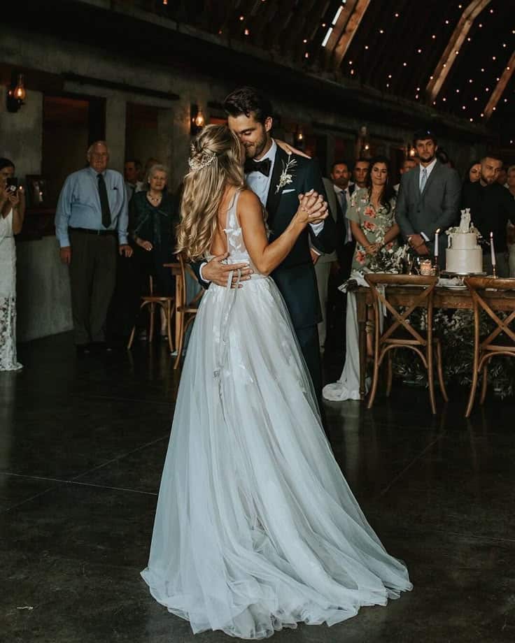 Bride and groom share their first dance at a rustic wedding reception in a warmly lit barn.