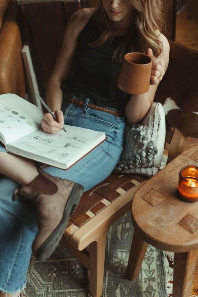 Woman sketching in a journal while sipping coffee, seated on a cozy chair with a candle nearby.