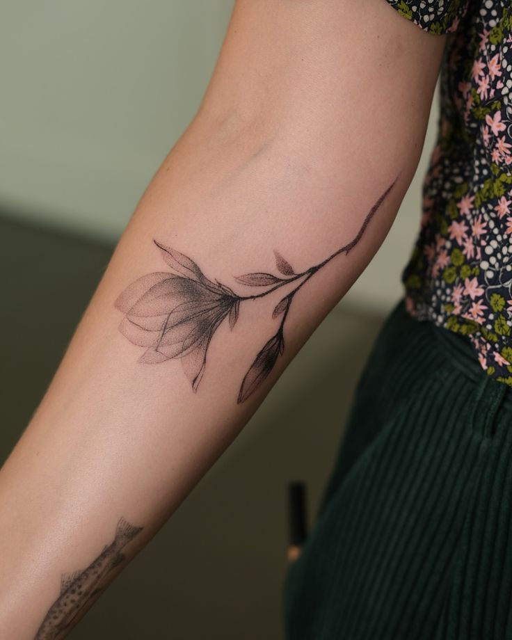 Minimalistic leaf branch tattoo on a person's forearm, complementing floral and textured clothing.
