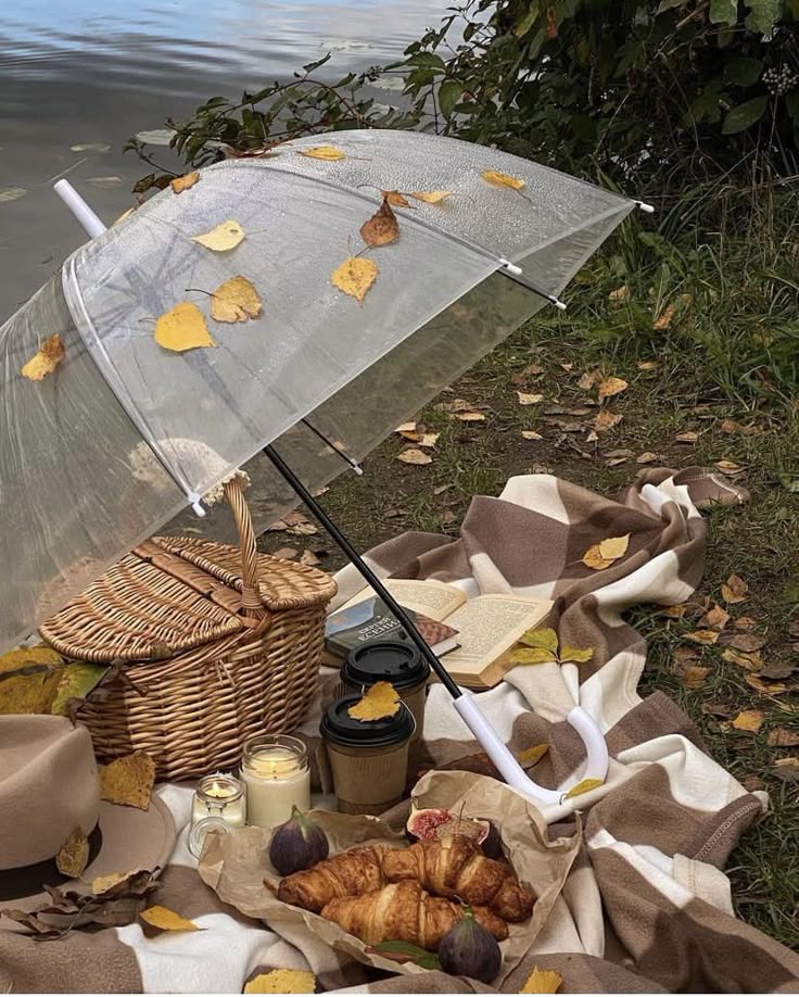 Autumn picnic scene with croissants, coffee, figs, candles, and a clear umbrella near a lake surrounded by leaves.