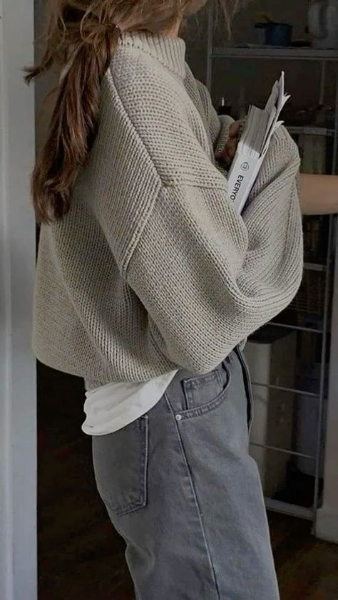 Woman in a cozy beige sweater holding documents, wearing jeans and standing sideways in a casual home setting.