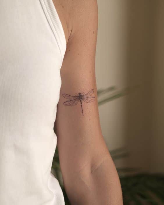 Delicate dragonfly tattoo on the upper arm of a person wearing a white tank top.