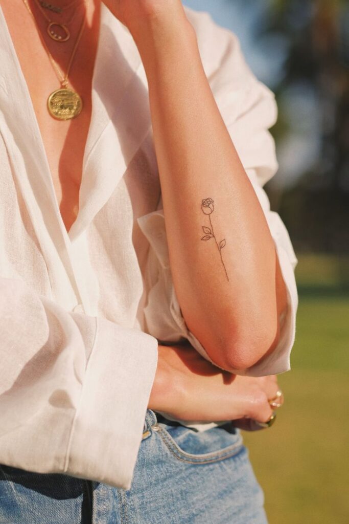 Minimal rose tattoo on forearm, woman in white blouse and jeans with gold necklace in sunny outdoor setting.