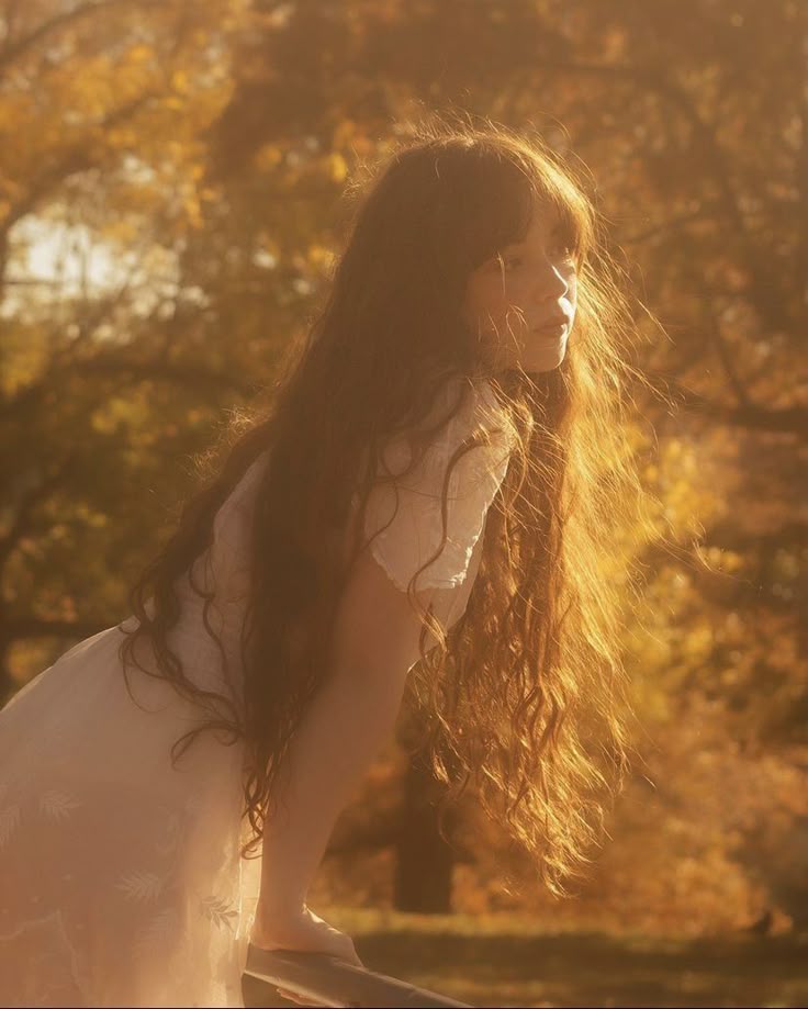 Young woman with long hair in autumn sunlight, wearing a white dress, surrounded by golden trees. Peaceful fall scenery.