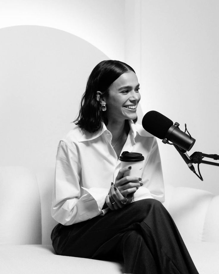 Woman smiling, holding a coffee cup, speaking into a microphone during a podcast recording session.