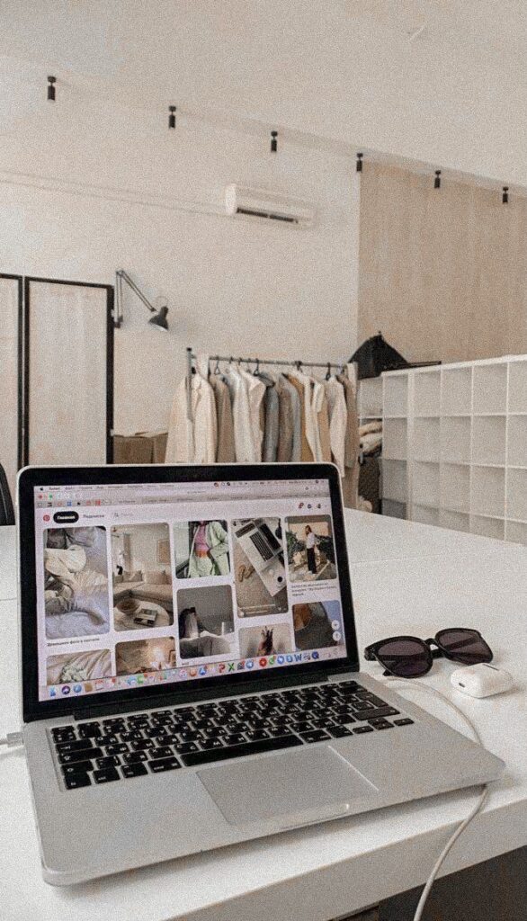 Laptop displaying Pinterest on table in minimalist office with clothing rack and sunglasses nearby.