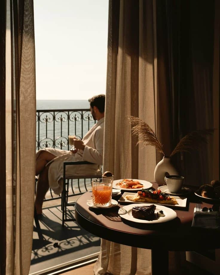Man in robe enjoys ocean view from balcony with breakfast table set for a luxurious morning.