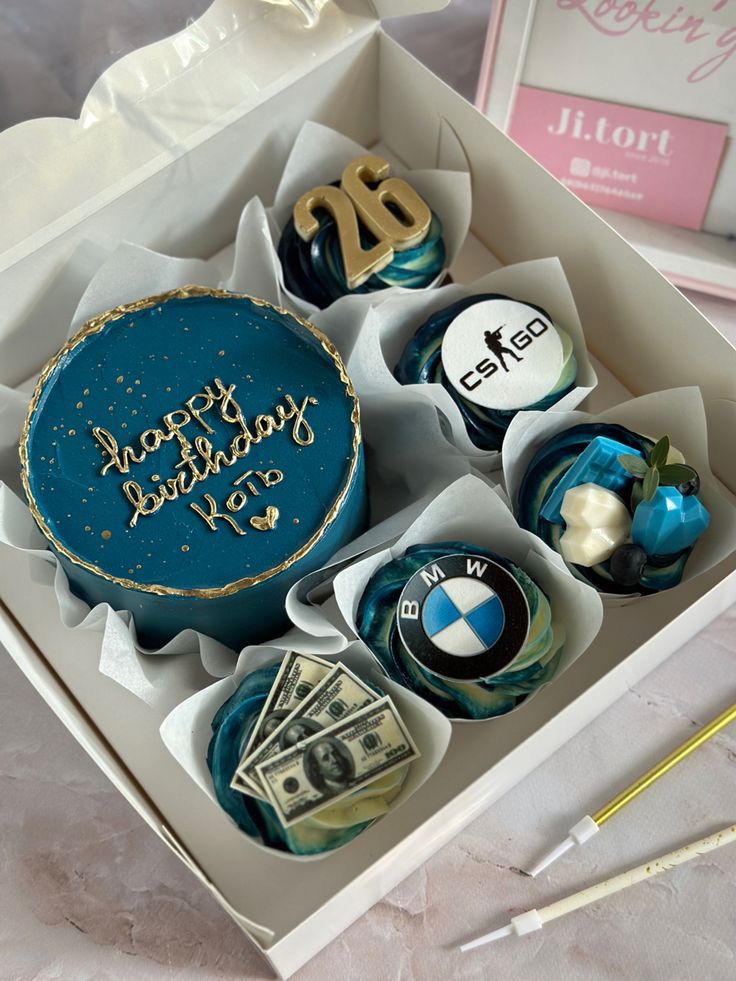 Box of themed birthday cakes with blue icing, featuring 26, BMW logo, and CSGO, alongside money decorations.