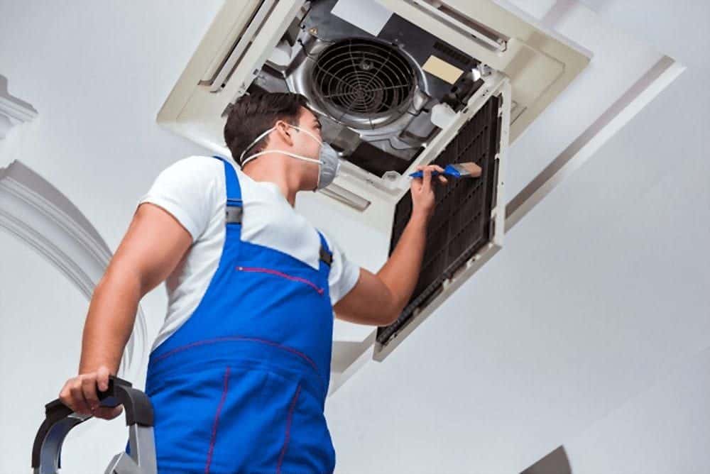 Technician cleaning and servicing an air conditioning unit on the ceiling, wearing blue overalls and a face mask.