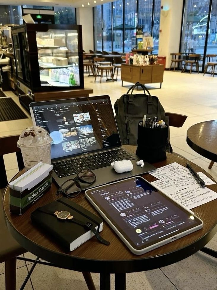 Cozy café workspace with a laptop, tablet, notebooks, and coffee on a round table, ideal for productivity.