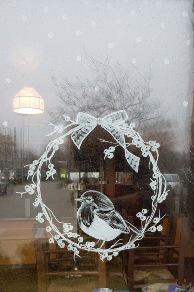 Bird illustration on window with a snowflake wreath and festive bow, reflecting a wintry street scene outside.