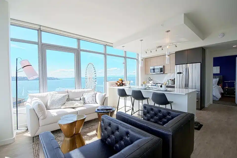 f7c6ec39ab9628e5b45c07b4c5ca156ac-f3497484155rd-w960_h720 Modern apartment living room with ocean view, sleek kitchen, and cozy seating area in neutral tones. | Sky Rye Design Modern apartment living room with ocean view, sleek kitchen, and cozy seating area in neutral tones.
