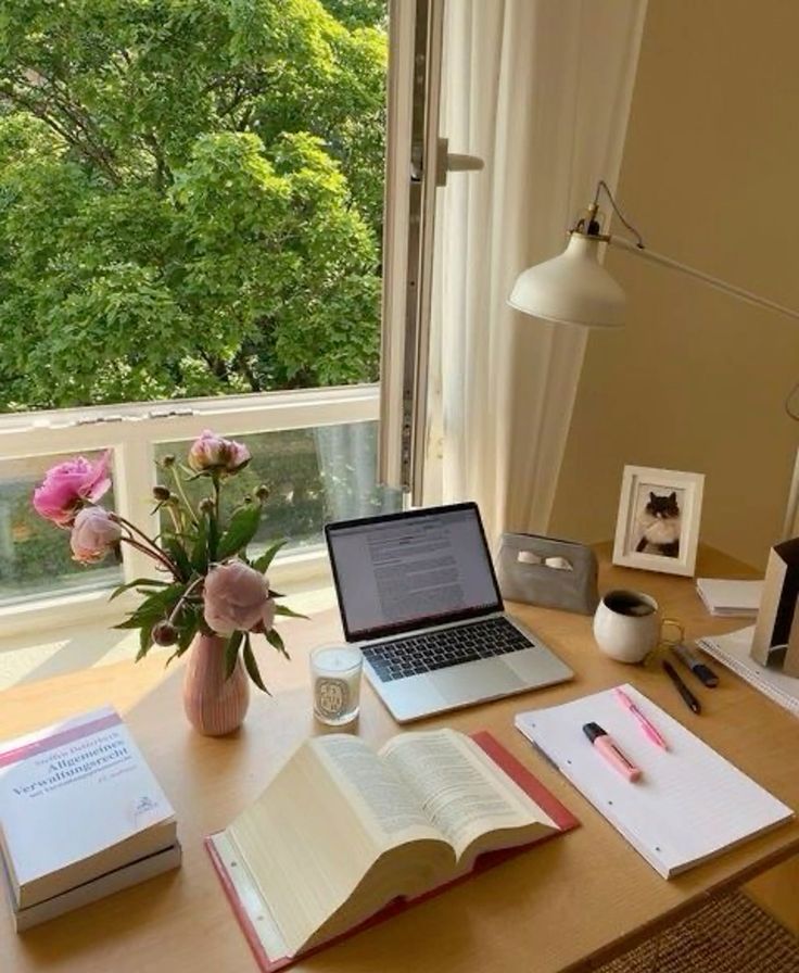 Cozy home office with laptop, open book, flowers, coffee, and notebook by a window with a green view.