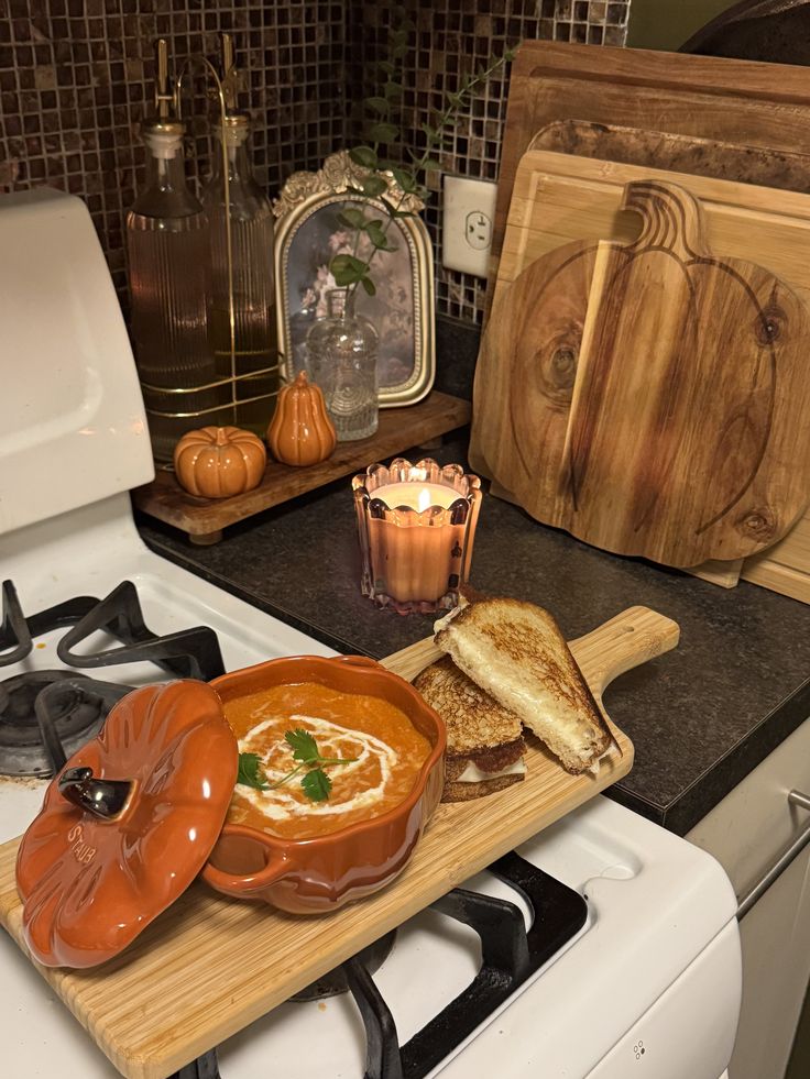 Pumpkin-shaped soup bowl with grilled cheese on stove, cozy fall kitchen decor with cutting boards and candle.