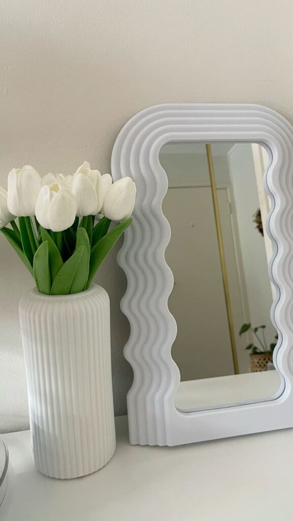 White tulips in a ribbed vase beside a wavy-edged mirror on a light-colored surface.