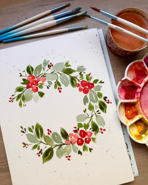 Watercolor floral wreath painting with red flowers and green leaves, surrounded by paintbrushes and a palette.
