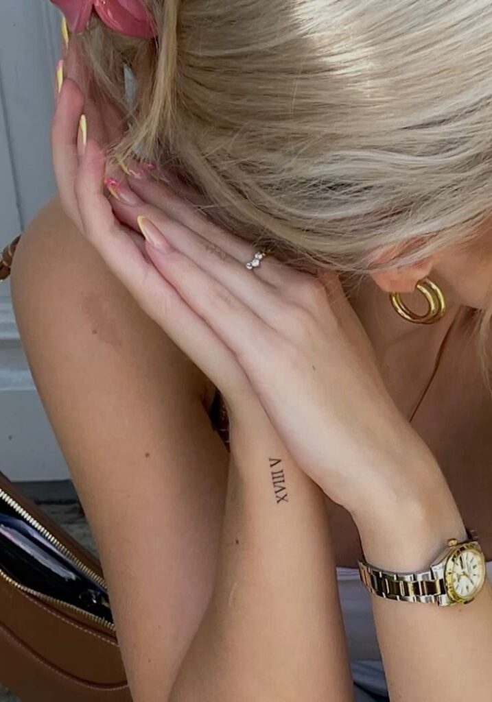 Close-up of a person with gold watch and hoop earrings, showcasing a small Roman numeral tattoo on their forearm.