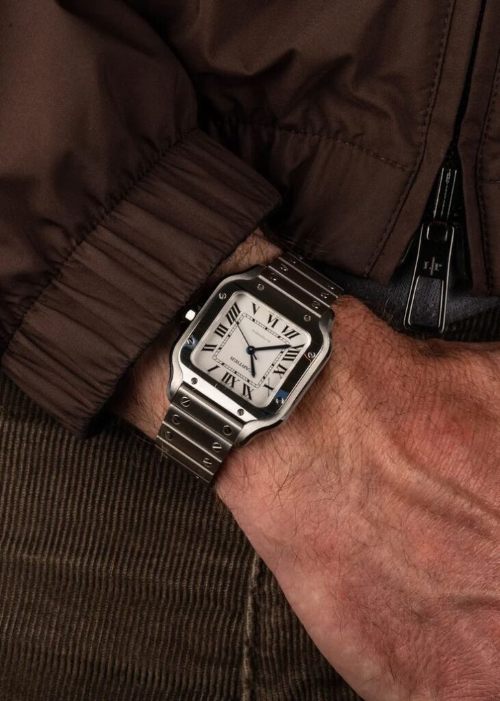 Man wearing luxury stainless steel wristwatch with Roman numerals on arm with brown jacket.